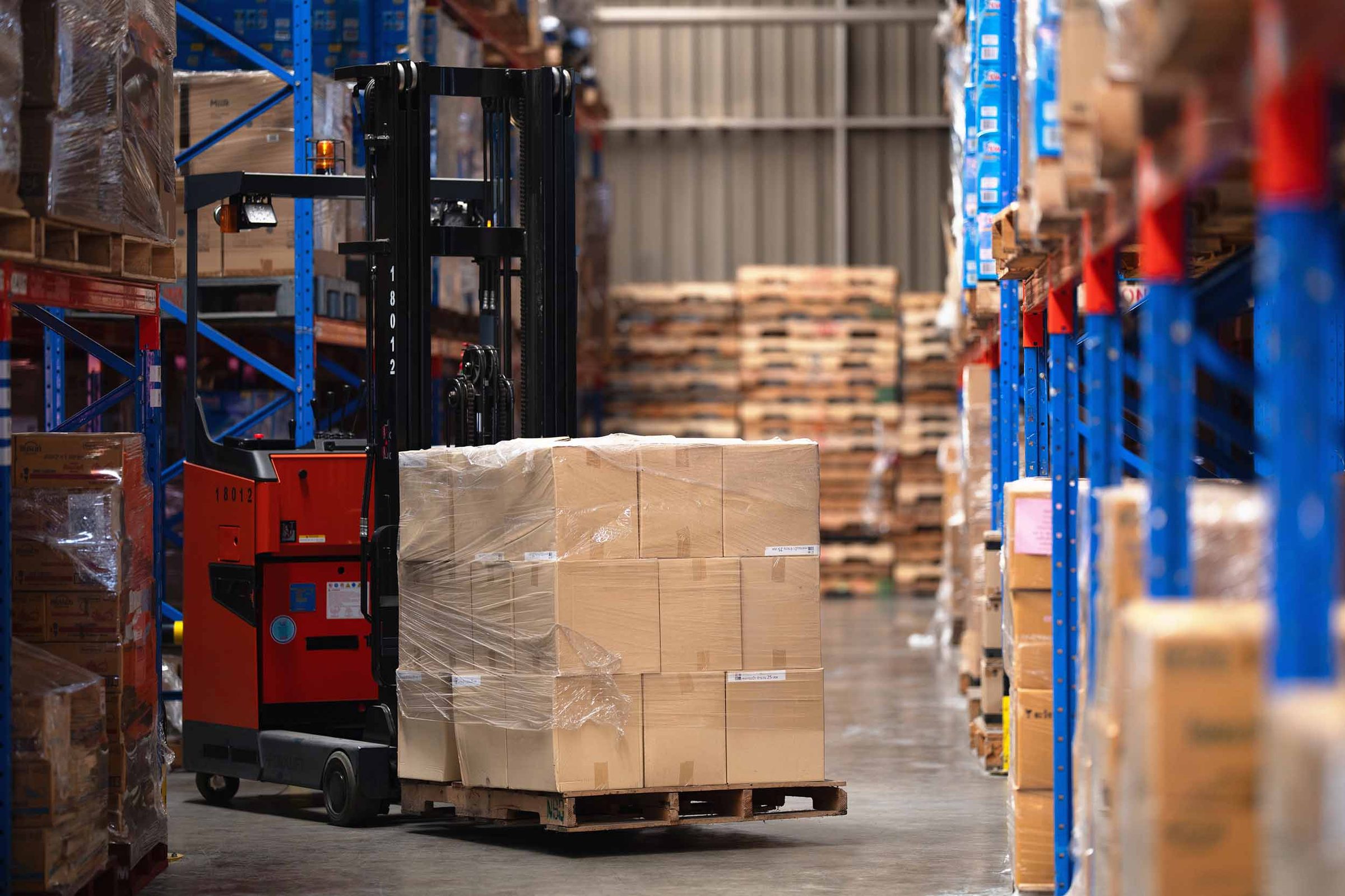 Wide shot of warehouse machinery handling palletised freight, with steel racking, wrapped loads and stored merchandise filling the frame on both sides of the storage aisle.